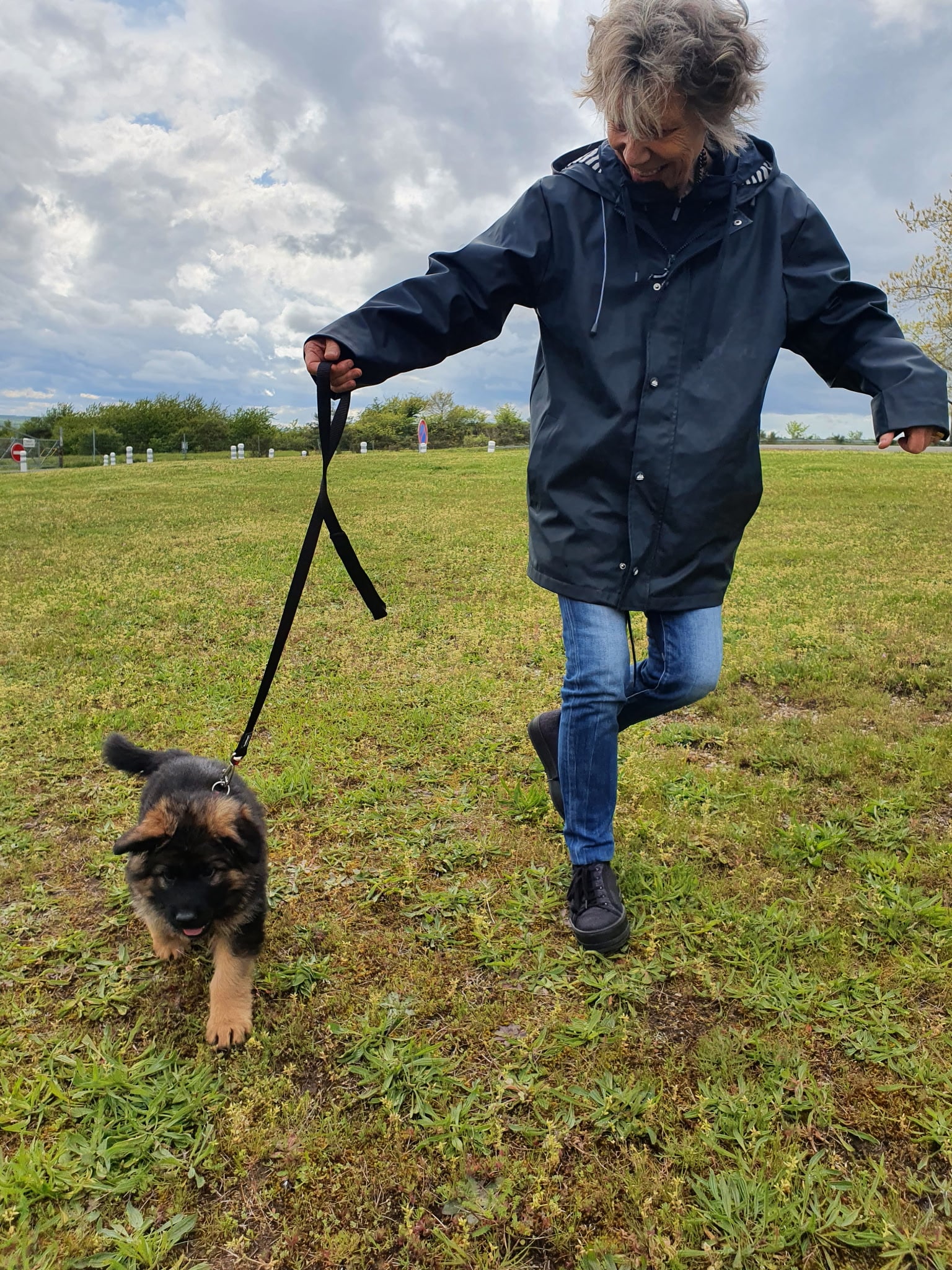 Femme joyeuse promenant un chiot Berger Allemand en laisse dans une prairie sous un ciel nuageux.